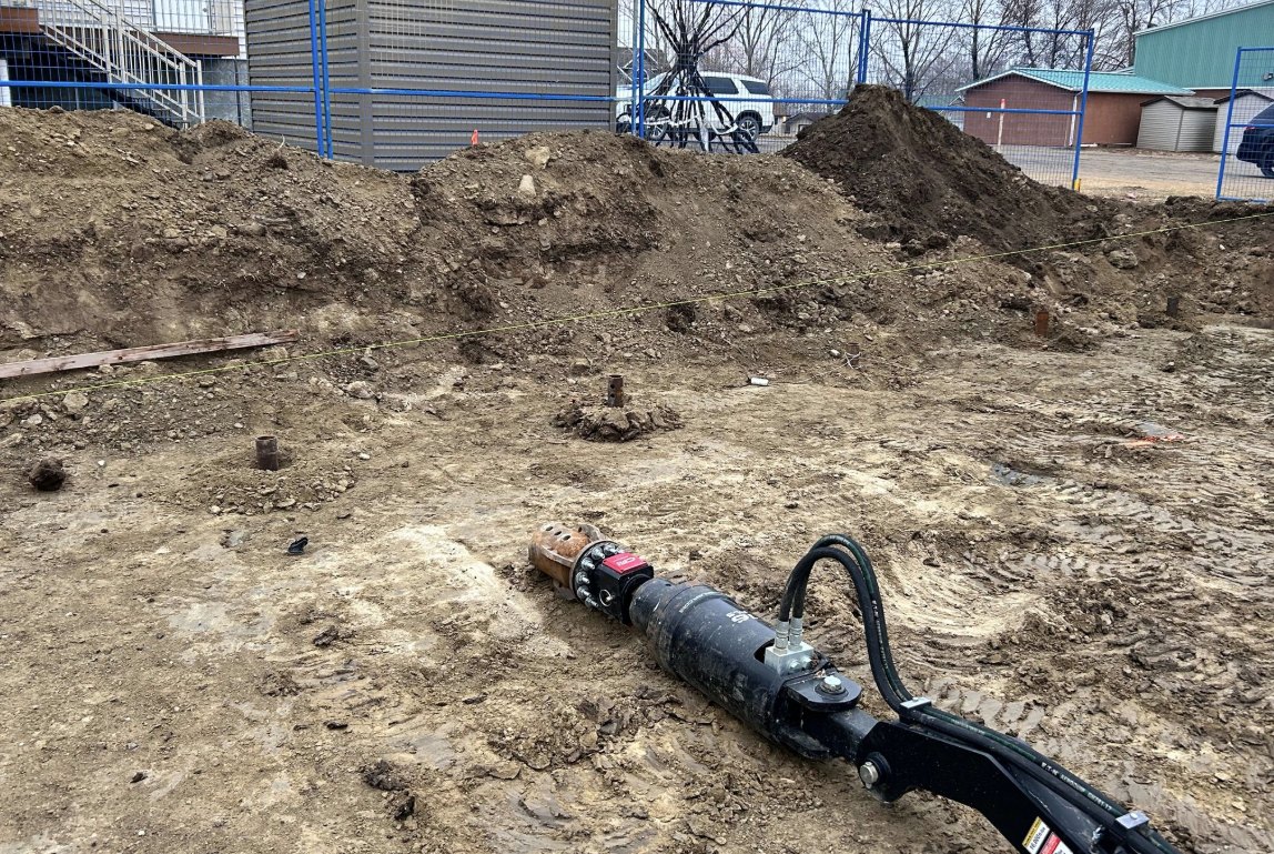A hydraulic torque drive head on an excavator beside helical piles already installed to grade on an excavated site